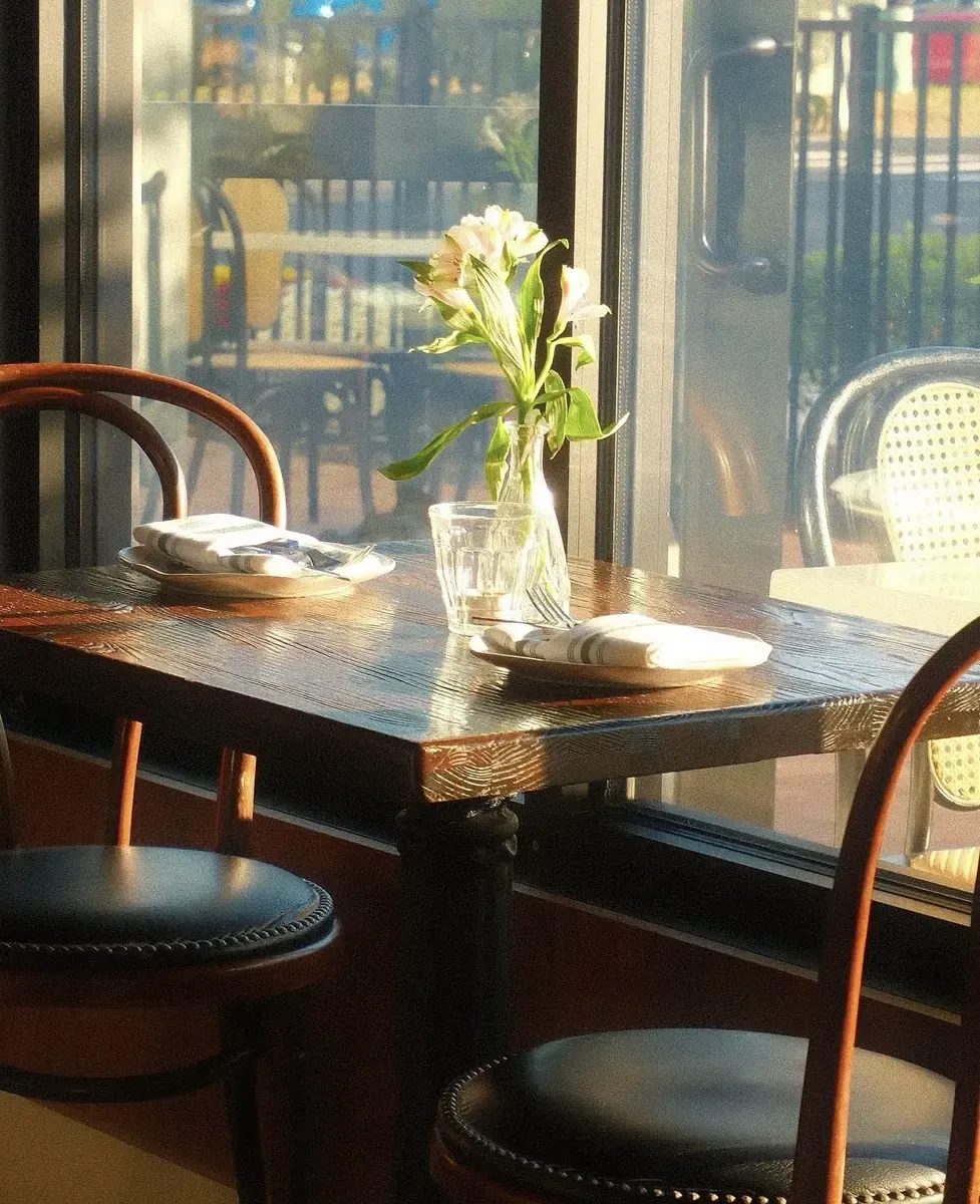 Bentwood bistro chairs at a window table inside Roto, with a small vase of flowers and low sun across the tabletop