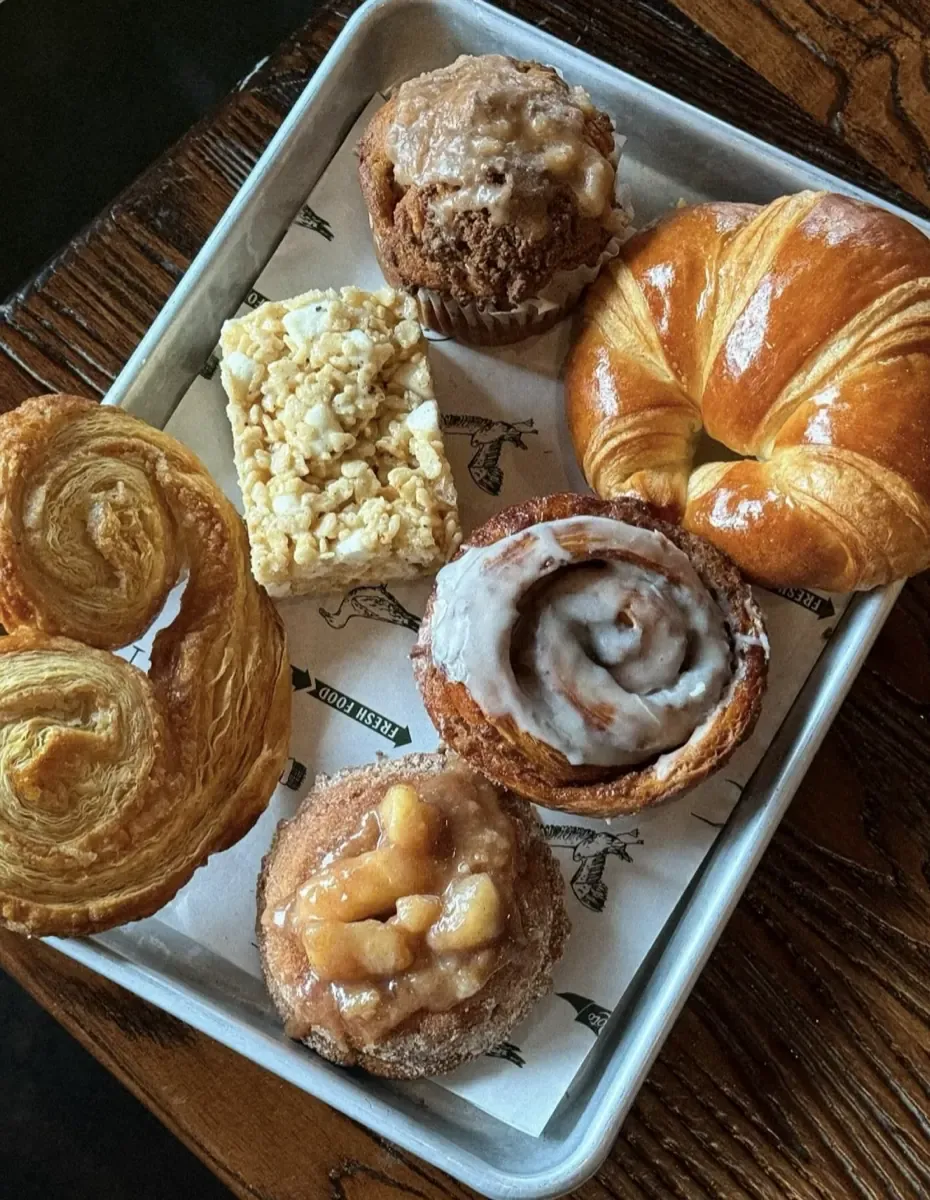 A metal tray of pastries on a wooden table: a croissant, an iced cinnamon roll, a laminated pastry, a crumb-topped muffin, a cereal treat square, and a doughnut topped with apple