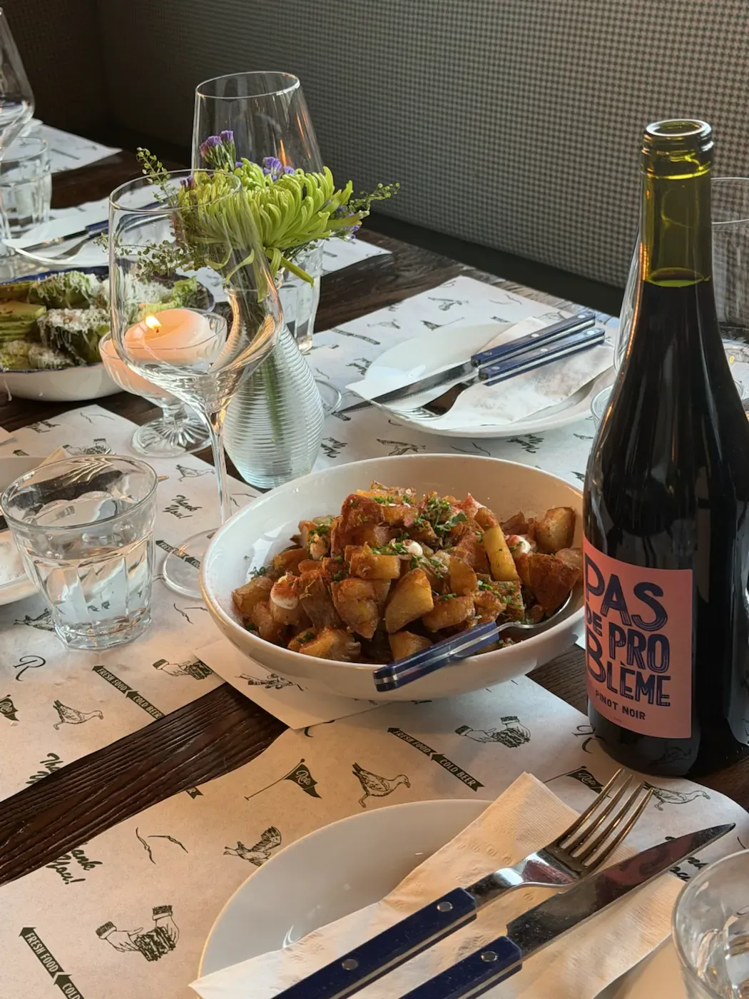 A table laid for dinner at Roto All Day, with a shared bowl, a bottle of wine, a lit candle and flowers