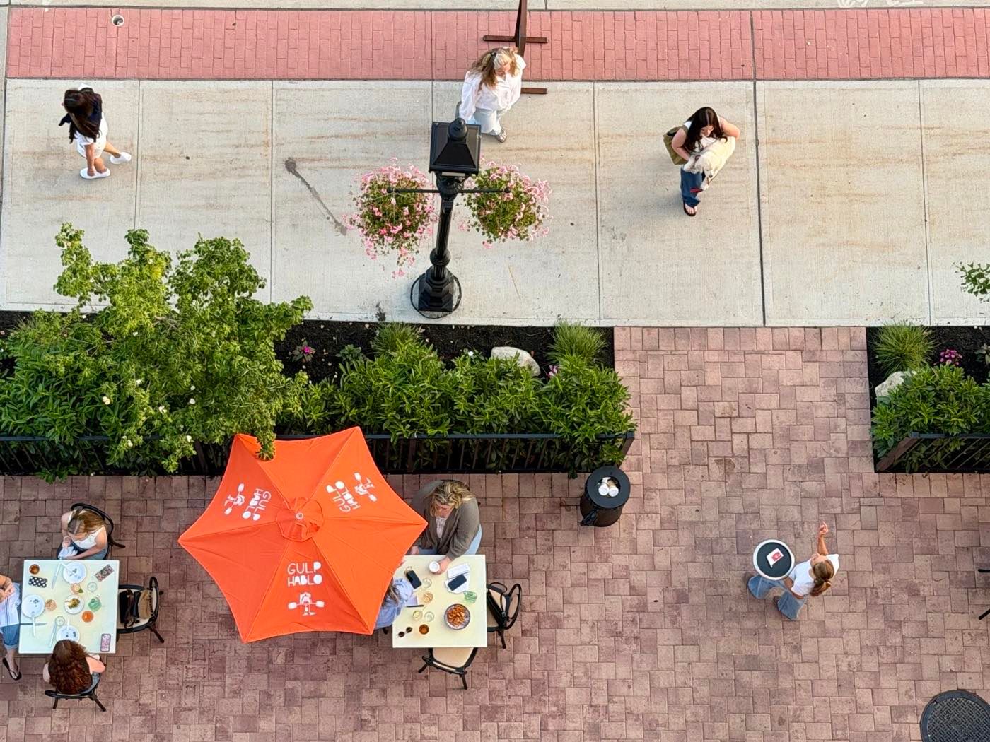 Overhead view of guests eating at sidewalk tables under an orange umbrella outside Roto All Day in Bay Shore